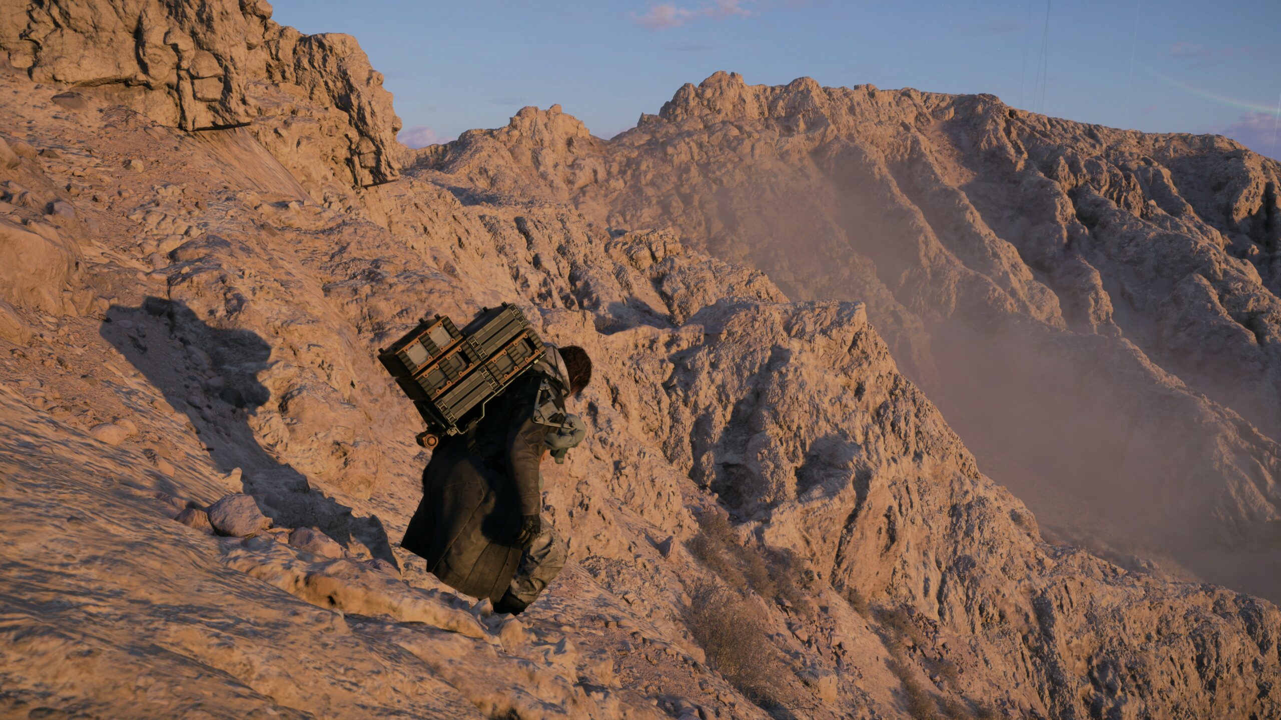 Hahmo kiipeää jyrkkää, hiekkaista ja kivikkoista rinteessä kantaen selässään suurta pinoa vihreitä ja ruskeita varustelaatikoita. Taustalla näkyy karu, auringonlaskun sävyttämä vuoristomaisema.