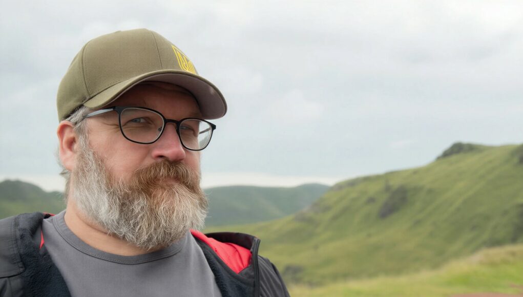 Lähikuva miehestä, jolla on harmaantunut parta, mustat silmälasit ja oliivinvihreä lippalakki, jossa on keltainen symboli. Hän katsoo suoraan kameraan rauhallinen ilme kasvoillaan. Taustalla näkyy sumuisia, vihreitä kukkuloita pilvisen taivaan alla.