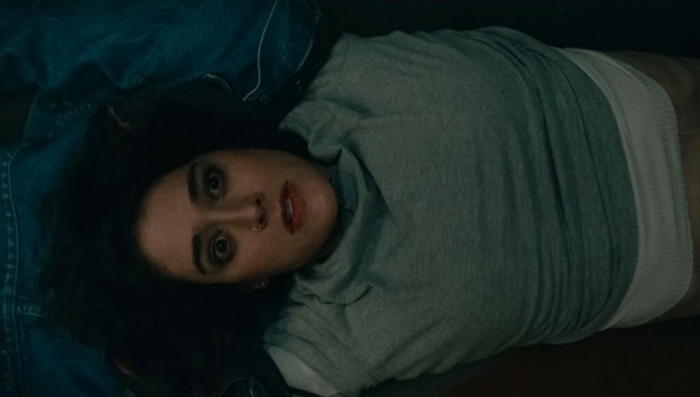 Ylhäältä päin kuvattu lähikuva naisesta, joka makaa selällään tummalla alustalla. Hänellä on päällään harmaa paita ja farkkutakki. Hänen silmänsä ovat ammollaan ja suu hieman raollaan, mikä välittää voimakkaan tunnetilan, kuten järkytyksen tai epäuskon. Kuva on valaistu hämärästi ja siinä on elokuvallinen tunnelma.