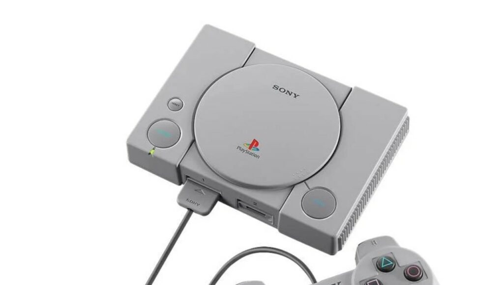Viistosta kuvattu alkuperäinen harmaa Sony PlayStation (PS1) -konsoli, jonka edessä on siihen kytketty klassinen ohjain. Konsolin päällä näkyvät pyöreä CD-aseman kansi, virta- ja avauspainikkeet sekä PlayStation-logo.