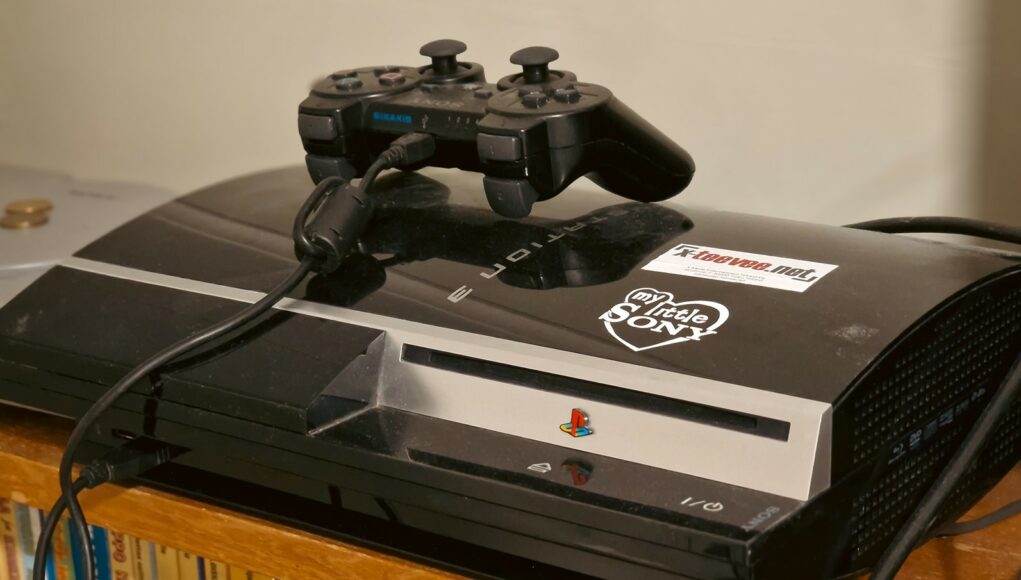 Alkuperäinen kiiltävän musta PlayStation 3 -konsoli, jonka päällä lepää musta langaton Sixaxis-ohjain. Konsolin kanteen on liimattu valkoinen "My Little Sony" -sydäntarra ja "teevee.net" -tarra. Konsoli sijaitsee puisella hyllyllä, jonka alla näkyy rivi kirjoja tai sarjakuvalehtiä.