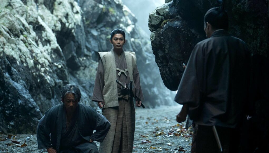 Kolme perinteisiin japanilaisiin asuihin pukeutunutta miestä hämärässä, kivikkoisessa poukamassa tai luolassa. Yksi mies seisoo keskellä tarkkaavaisena katana vyöllään, toinen on kyykistynyt maahan hänen vierellään, ja kolmas hahmo näkyy selin etualalla oikealla.