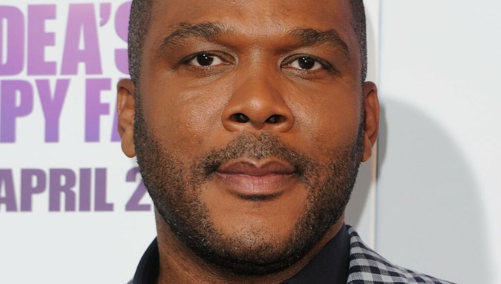 tyler-perry Lähikuva Tyler Perrystä katsomassa suoraan kameraan vakavalla ilmeellä. Hänellä on lyhyt sänki ja yllään tummansininen kauluspaita sekä harmaa-valkoruudullinen pikkutakki. Taustalla näkyy osa elokuvajulisteesta, jossa on purppuraa tekstiä.