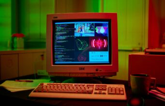 Pelialalla käytössä yli 500 tekoälysovellusta, joista alle tusina on oikeasti hyödyllisiä Tunnelmallinen kuva vanhasta IBM-tietokoneesta, joka on asetettu työpöydälle hämärässä huoneessa. Huone on valaistu voimakkailla vihreillä ja punaisilla neonvaloilla. Tietokoneen näytöllä näkyy koodia sekä erilaisia graafisia elementtejä ja kuvakaappauksia. Pöydällä on myös näppäimistö, hiiri, kynä, paperipino ja keraaminen muki. Tietokoneen keskusyksikkö näkyy osittain näytön oikealla puolella.