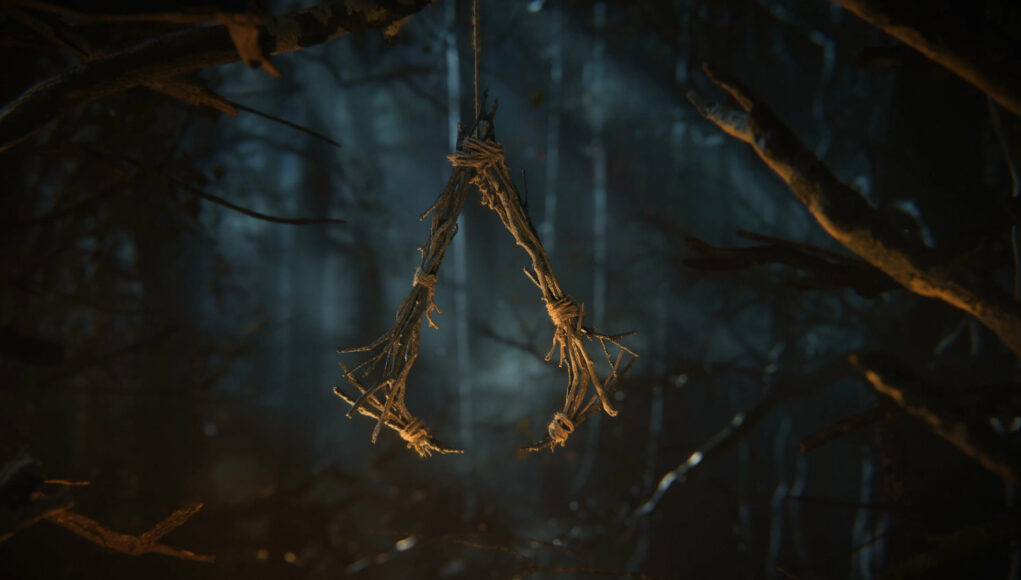 Synkkä ja tunnelmallinen kuva, jossa oksista ja narusta kyhätty kolmionmuotoinen symboli roikkuu ilmassa puiden siimeksessä. Symboli muistuttaa Assassin’s Creed -pelisarjan logoa, mutta se on tehty karkeasti luonnonmateriaaleista. Taustalla näkyy pimeä, sumuinen metsä, jota valaisee heikko, kylmä valo. Etualalla on epätarkkoja oksia, ja tunnelma on mystinen ja hieman pahaenteinen.