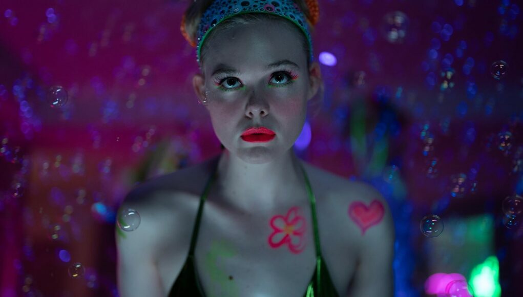 Lähikuva naisesta (Elle Fanning), jolla on kalpeat kasvot ja kirkkaan vaaleanpunainen huulipuna. Hän katsoo suoraan kameraan intensiivisellä ilmeellä. Hänen vartaloonsa ja kasvoihinsa on maalattu neonvärisiä kuvioita, kuten sydän ja kukka, ja hänen silmämeikkinsä on neonvihreä. Hänellä on päässään kimaltelevan sininen panta. Tausta on hämärä ja täynnä vaaleanpunaisia neonvaloja sekä leijuvia saippuakuplia, jotka luovat unenomaisen tunnelman.