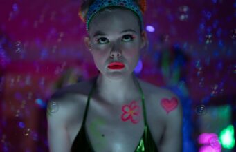 Käyttikö Elle Fanning vartalostunttia uuden sarjansa alastonkohtauksissa? Lähikuva naisesta (Elle Fanning), jolla on kalpeat kasvot ja kirkkaan vaaleanpunainen huulipuna. Hän katsoo suoraan kameraan intensiivisellä ilmeellä. Hänen vartaloonsa ja kasvoihinsa on maalattu neonvärisiä kuvioita, kuten sydän ja kukka, ja hänen silmämeikkinsä on neonvihreä. Hänellä on päässään kimaltelevan sininen panta. Tausta on hämärä ja täynnä vaaleanpunaisia neonvaloja sekä leijuvia saippuakuplia, jotka luovat unenomaisen tunnelman.