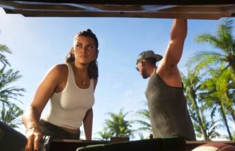 5 vuotta GTA 6 -peliä kehittänyt devaaja kertoo podcastissa, mitä tietää tulevasta hittipelistä Alaviistosta otettu kuva auton tavaratilasta, josta katsotaan ylöspäin kohti kahta hahmoa, Luciaa ja Jasonia. Lucia seisoo vasemmalla valkoisessa hihattomassa paidassa ja katsoo vakavana sivulle, samalla kun hänen kätensä lepää tavaratilan reunalla. Jason seisoo oikealla harmaassa hihattomassa paidassa ja lippalakissa, kurkistaen sisään autoon. Taustalla näkyy kirkas sininen taivas ja useita palmuja.