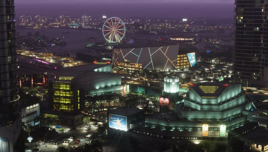 Laaja lintuperspektiivinäkymä vilkkaasta Vice City -suurkaupungista iltahämärässä. Etualalla on moderneja rakennuksia, kuten konserttitalo ja areena, jonka julkisivua koristavat neonvalot. Taustalla näkyy valaistu maailmanpyörä rannassa, satama-alueen nostureita ja silta, jolla kulkee autoja. Taivas on sävyltään syvän purppura.