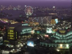 Julkaistaanko Grand Theft Auto VI:n odotettu kolmas traileri huhtikuussa? Laaja lintuperspektiivinäkymä vilkkaasta Vice City -suurkaupungista iltahämärässä. Etualalla on moderneja rakennuksia, kuten konserttitalo ja areena, jonka julkisivua koristavat neonvalot. Taustalla näkyy valaistu maailmanpyörä rannassa, satama-alueen nostureita ja silta, jolla kulkee autoja. Taivas on sävyltään syvän purppura.