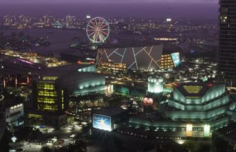 Julkaistaanko Grand Theft Auto VI:n odotettu kolmas traileri huhtikuussa? Laaja lintuperspektiivinäkymä vilkkaasta Vice City -suurkaupungista iltahämärässä. Etualalla on moderneja rakennuksia, kuten konserttitalo ja areena, jonka julkisivua koristavat neonvalot. Taustalla näkyy valaistu maailmanpyörä rannassa, satama-alueen nostureita ja silta, jolla kulkee autoja. Taivas on sävyltään syvän purppura.