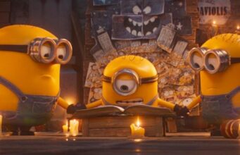 Kätyrit palaavat valkokankaille uudessa Minions & Monsters -elokuvassa Kolme Kätyrit-hahmoa (Minions) seisoo hämärässä huoneessa pöydän äärellä, jolla on avoin kirja. Keskimmäinen, yksisilmäinen Kätyri lukee kirjaa keskittyneenä, kun taas kaksi muuta pitelevät toisiaan käsistä hänen sivuillaan. Pöydällä ja lattialla palaa useita kynttilöitä, jotka luovat lämmintä valoa. Taustalla näkyy seinä, joka on täynnä piirustuksia ja luonnoiteltuja hahmoja.