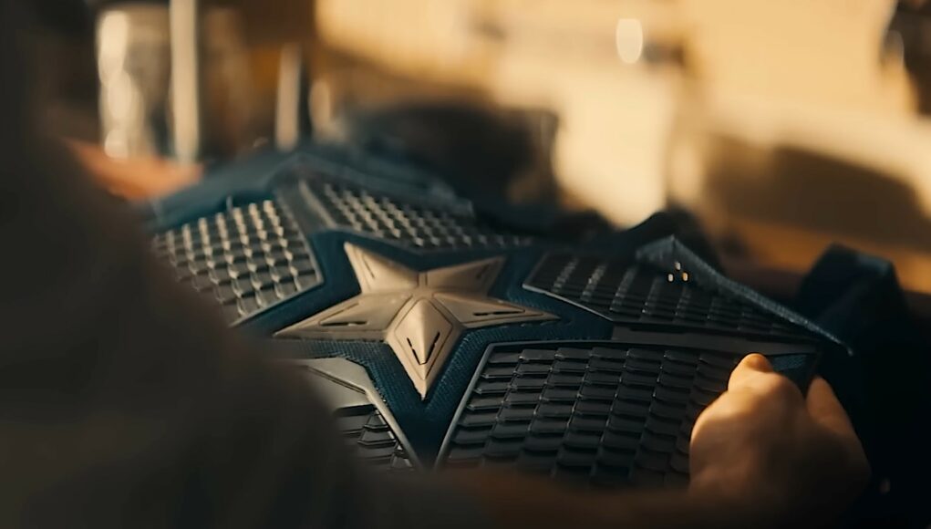 Lähikuva Captain American univormun rintaosasta, jota joku pitelee käsissään. Puvun keskellä on suuri, metallinen tähtisymboli, ja sitä ympäröi tummansininen, suomumainen ja tekninen kangasrakenne. Valaistus on lämmin ja hämärä, ja tausta on sumennettu, mikä korostaa puvun yksityiskohtia ja materiaalin tuntua.