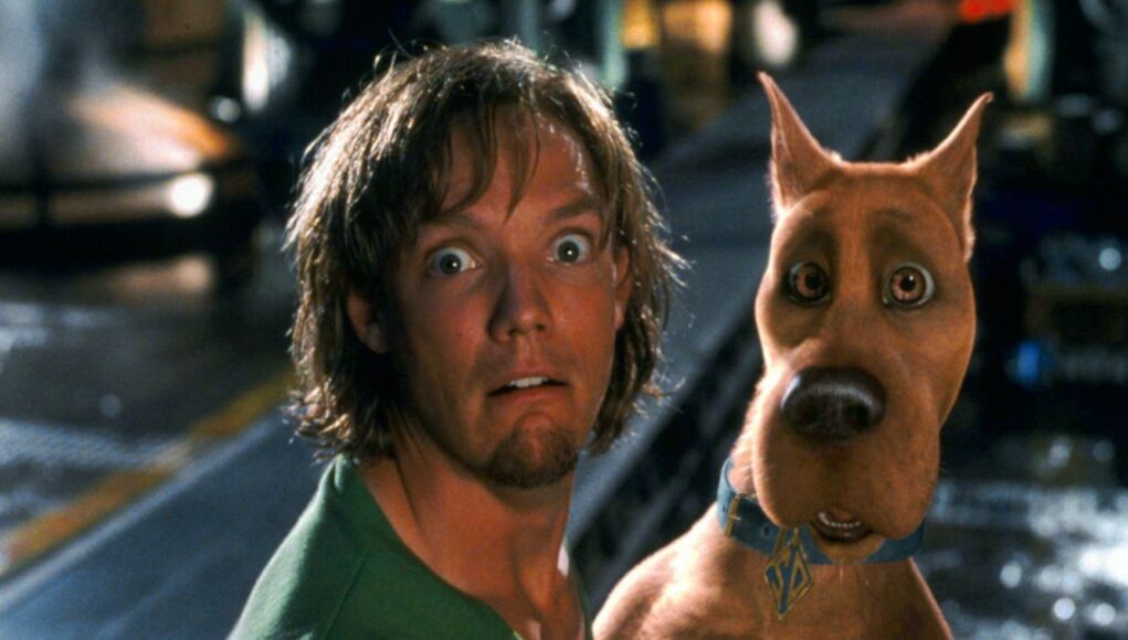 Lähikuva elokuvasta Scooby-Doo. Kuvassa Shaggy (Matthew Lillard) ja tietokoneanimoitu Scooby-Doo-koira katsovat suoraan kameraan kauhistuneet ja järkyttyneet ilmeet kasvoillaan. Shaggylla on yllään tunnusomainen vihreä t-paitansa, ja Scoobylla on sininen kaulapanta, jossa on kultainen SD-nimilaatta. Tausta on hämärä ja hieman epätarkka, muistuttaen tehdas- tai varastoympäristöä.