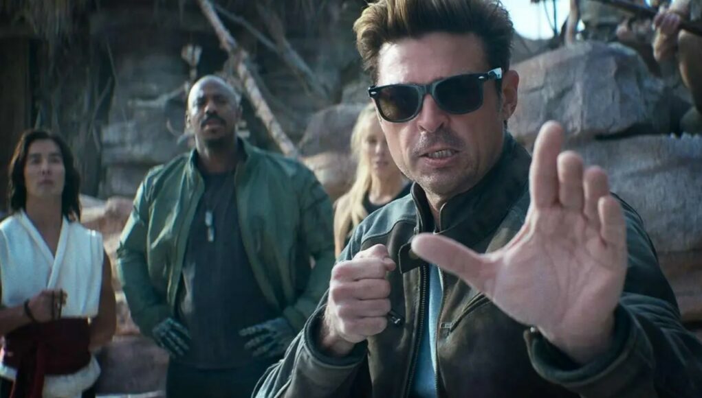 Elokuvallinen kohtaus, jossa aurinkolaseihin ja ruskeaan nahkatakkiin pukeutunut mies (Johnny Cage) on etualalla taisteluasennossa, kämmen ojennossa kohti kameraa. Hänen takanaan seisoo muita hahmoja, muun muassa vihreään takkiin pukeutunut mies (Jax) ja valkoiseen asuun pukeutunut mies (Liu Kang). Taustalla näkyy karu, kallioinen ympäristö.