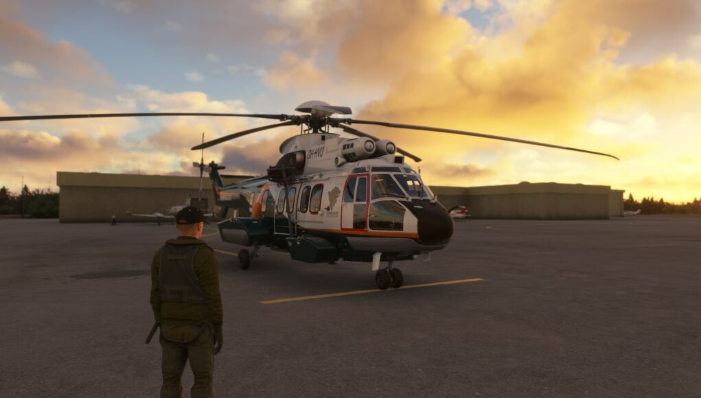 Pelinäkymä Microsoft Flight Simulator -pelistä, jossa lentäjä seisoo selin katsojaan lentokentän asfaltilla katsoen Rajavartiolaitoksen Super Puma -helikopteria. Helikopteri on väritykseltään valkoinen ja tummanvihreä oransseilla yksityiskohdilla, ja siinä on rekisteritunnus OH-HVQ. Taustalla näkyy matalia lentokenttärakennuksia ja upea ilta-aurinko, joka värjää pilvet kullankeltaisiksi ja oransseiksi.