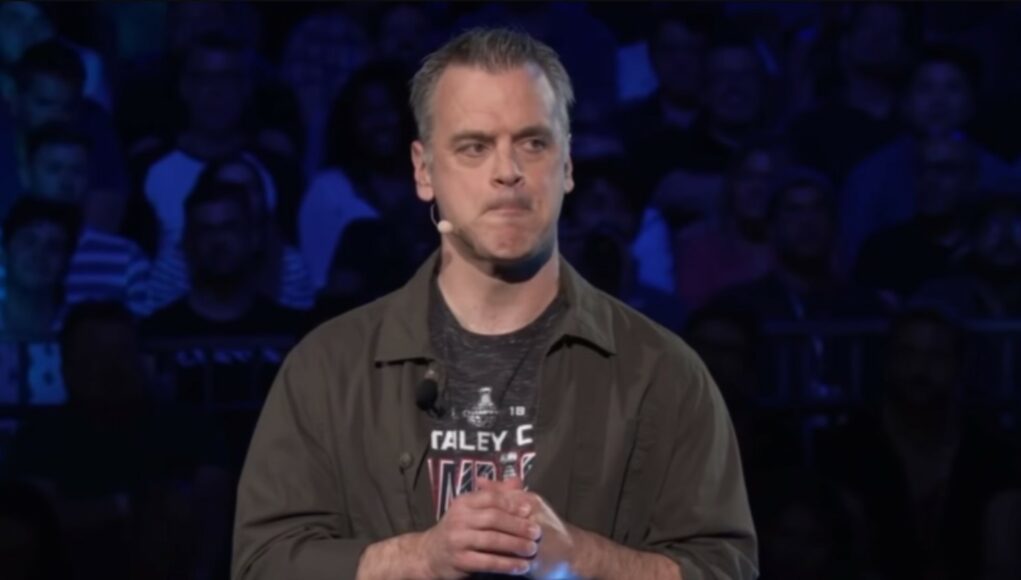 Keski-ikäinen mies, jolla on lyhyet harmahtavat hiukset, seisoo lavalla suuren yleisön edessä. Hänellä on yllään harmaa t-paita ja sen päällä avoin tummanvihreä kauluspaita. Miehellä on päässään pieni sankamikrofoni, ja hän puristaa huuliaan yhteen vakavalla ilmeellä kädet ristissä vatsan korkeudella. Taustalla näkyy hämärästi suuri määrä ihmisiä katsomossa.