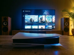 Oletko tiennyt tätä ominaisuutta PS5:ssä? Tunnelmallinen, 35 mm:n filmikameralla otettu valokuva olohuoneesta iltahämärässä. Etualalla puisella pöydällä lepää PlayStation 5 -konsoli, jonka sininen merkkivalo hohtaa pimeässä. Taustalla on suuri televisio, jossa näkyy PlayStation Store -valikko pelikuvakkeineen. Television molemmin puolin on puiset lattiakaiuttimet, ja oikeassa reunassa on viherkasvi, jota valaisee lämmin, keltainen tunnelmavalo. Huoneen valaistus on hämärä ja koostuu television sinertävästä kajosta sekä nurkkauksen pehmeästä keltaisesta valosta.