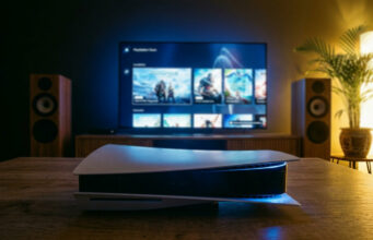 Oletko tiennyt tätä ominaisuutta PS5:ssä? Tunnelmallinen, 35 mm:n filmikameralla otettu valokuva olohuoneesta iltahämärässä. Etualalla puisella pöydällä lepää PlayStation 5 -konsoli, jonka sininen merkkivalo hohtaa pimeässä. Taustalla on suuri televisio, jossa näkyy PlayStation Store -valikko pelikuvakkeineen. Television molemmin puolin on puiset lattiakaiuttimet, ja oikeassa reunassa on viherkasvi, jota valaisee lämmin, keltainen tunnelmavalo. Huoneen valaistus on hämärä ja koostuu television sinertävästä kajosta sekä nurkkauksen pehmeästä keltaisesta valosta.