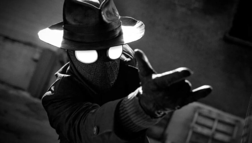 Mustavalkoinen kuva Spider-Man Noir -hahmosta, joka on pukeutunut maskiin, fedora-hattuun ja trenssitakkiin. Hahmon suuret, pyöreät silmälasit hohdavat kirkkaan valkoisina muuten tummassa kuvassa. Spider-Man Noir ojentaa käsinettä pitelevää kättään kohti katsojaa Spider-Manille tyypillisessä seittien ammunta-asennossa. Taustalla näkyy hämärä kaupunkiympäristö.