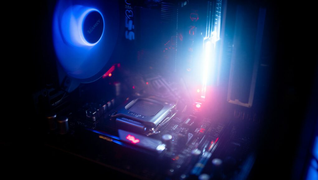 Lähikuva tietokoneen sisälmystä ja emolevystä, joka on valaistu näyttävillä sinisillä ja punaisilla LED-valoilla. Vasemmassa yläkulmassa näkyy kirkas sininen tuuletin, ja oikealla puolella hohtaa voimakas valonlähde muistikampojen kohdalla. Keskellä näkyy prosessorikanta ja ympärillä emolevyn komponentteja, kuten kondensaattoreita ja piirejä, hämärässä valaistuksessa.