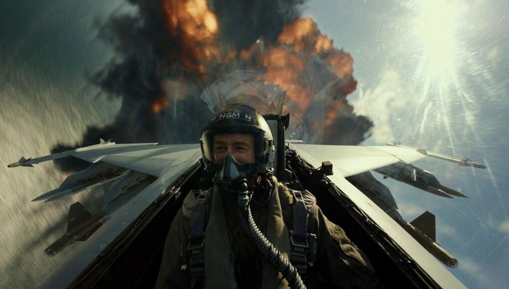 Toiminnallinen elokuvakohtaus hävittäjän ohjaamosta käsin kuvattuna. Lentäjä Hangman (Glen Powell) istuu ohjaamossa täydessä varustuksessa, kypärä ja happimaski kasvoillaan, ja katsoo sivuun hymyillen. Aivan hävittäjän pyrstön takana näkyy massiivinen räjähdys, josta nousee mustaa savua ja oransseja liekkejä. Kirkas aurinko paistaa oikealta ylhäältä, luoden voimakkaita valonheijastuksia linssiin.