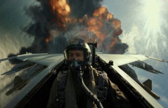 Top Gun 3 menee tuotantoon, mutta sen on vaikea lunastaa Top Gun: Maverickin menestys Toiminnallinen elokuvakohtaus hävittäjän ohjaamosta käsin kuvattuna. Lentäjä Hangman (Glen Powell) istuu ohjaamossa täydessä varustuksessa, kypärä ja happimaski kasvoillaan, ja katsoo sivuun hymyillen. Aivan hävittäjän pyrstön takana näkyy massiivinen räjähdys, josta nousee mustaa savua ja oransseja liekkejä. Kirkas aurinko paistaa oikealta ylhäältä, luoden voimakkaita valonheijastuksia linssiin.