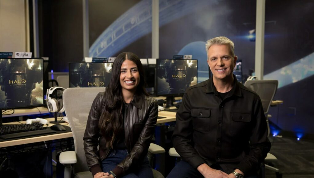 Kaksi ihmistä, nainen ja mies, istuvat hymyillen toimistomaisessa studiossa. Naisella on pitkät tummat hiukset ja ruskea nahkatakki, ja miehellä on harmaat hiukset ja musta kauluspaita. Heidän taustallaan on useita tietokonenäyttöjä, joissa näkyy Halo-pelin logo ja Master Chief -hahmo. Taustaseinällä on suuri graafinen kuva Halo-pelin rengasmaailmasta. Huoneessa on ammattimainen valaistus, jossa on sinisiä korostusvaloja lattianrajassa.