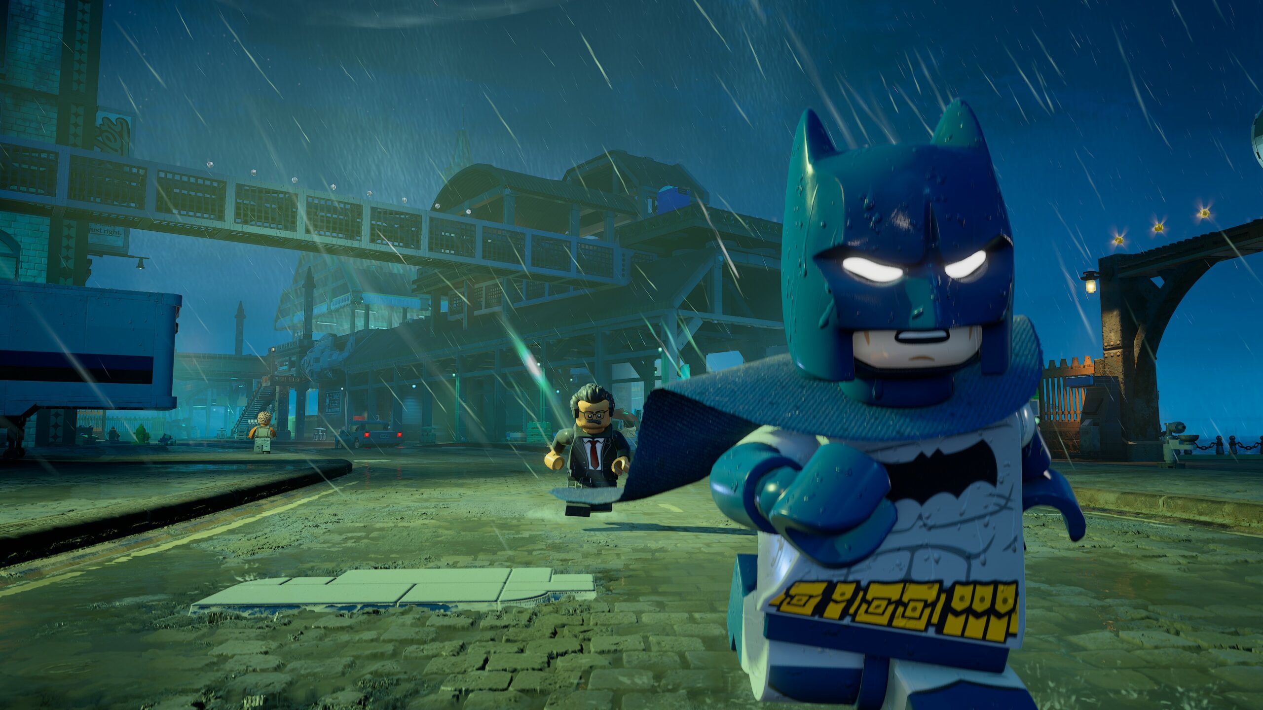 Kuvakaappaus videopelistä, jossa Lego Batman juoksee etualalla kohti kameraa kaatosateessa. Hahmolla on yllään harmaa puku sinisellä naamiolla ja viitalla, ja hänen silmänsä loistavat valkoisina. Taustalla näkyy sateen piiskaama satamalaituri tai katu, jossa Batmanin perässä juoksee pukuun pukeutunut vihainen Lego-hahmo (mahdollisesti komisario Gordon tai etsivä). Kauempana häämöttää teollisuusrakennuksia ja silta yöllisessä, sinisessä valaistuksessa.