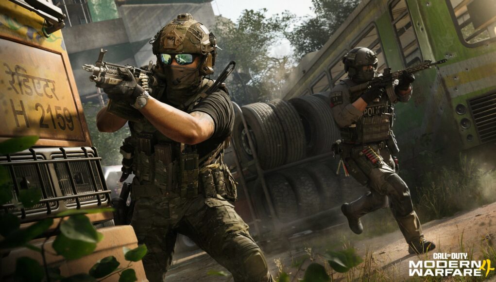 Toiminnallinen julkaisukuva Call of Duty: Modern Warfare III -videopelistä, jossa kaksi raskaasti varustautunutta sotilasta on taistelutilanteessa urbaanissa, hylätyssä ympäristössä päivänvalossa. Etualalla vasemmalla oleva sotilas tähtää konekiväärillä suoraan eteenpäin; hänellä on yllään maastokuvioinen kypärä, aurinkolasit, kasvomaski ja taktinen liivi täynnä varusteita. Oikealla puolella taustempana toinen sotilas, jolla on "POLICE"-tekstillä varustettu liivi, etenee juosten rynnäkkökivääri valmiina. Heidän välissään on kuorma-auton lava täynnä suuria renkaita, ja oikealla näkyy vihreän, hylätyn bussin kylki. Kuvan oikeassa alakulmassa on pelin virallinen logo "CALL OF DUTY MODERN WARFARE 4".