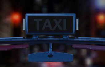 Sega julkisti uuden Crazy Taxi -pelin? Lähikuva taksin katolla olevasta valokyltistä yöaikaan. Kyltti on suorakaiteen muotoinen, tumma ja siinä lukee selkeällä fontilla sana "TAXI". Kuva on otettu auton sisältä tai aivan tuulilasin tasolta, sillä kyltin alapuolella näkyy osa auton korirakennetta ja taustapeili. Tausta on erittäin epätarkka (bokeh-efekti), ja siinä siintää kaupungin iltavaloja sekä punaista hehkua, mikä luo kuvaan urbaanin ja elokuvallisen tunnelman.
