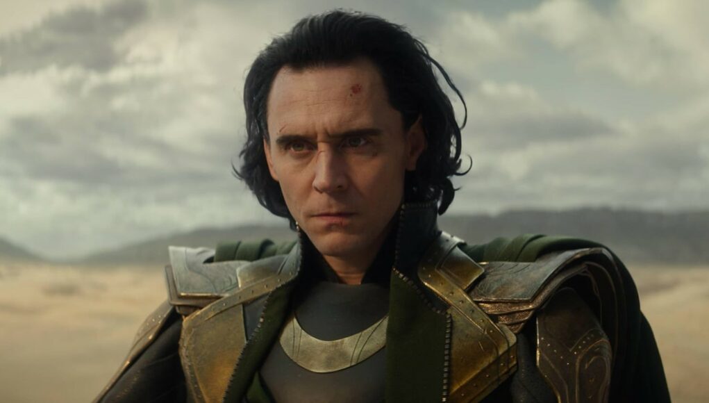 Lähikuva Tom Hiddlestonin esittämästä Loki-hahmosta Marvel-elokuvasta. Lokilla on yllään yksityiskohtainen vihreä ja kultainen asgardilainen haarniska. Hänen mustat hiuksensa ovat sekaisin, ja hänen kasvoillaan on pieniä naarmuja ja verta, mikä viittaa juuri päättyneeseen taisteluun. Lokilla on vakava ja päättäväinen ilme. Taustalla näkyy hämärä, autio aavikkomaisema pilvisen taivaan alla.