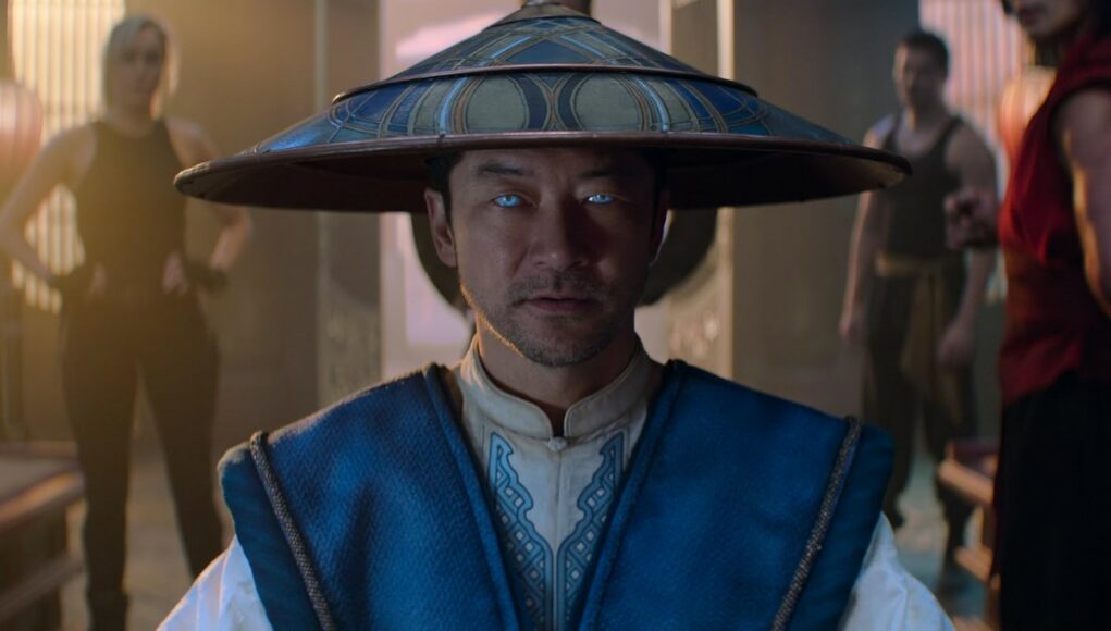 Lähikuva Mortal Kombat -elokuvan Lord Raiden -hahmosta. Hänellä on päässään perinteinen leveälierinen aasialainen hattu ja yllään sinivalkoinen asu. Hahmon silmät hehkuvat kirkkaan vaaleansinisinä, mikä viittaa yliluonnollisiin voimiin. Taustalla näkyy epätarkasti muita hahmoja, kuten Sonya Blade ja Liu Kang, perinteisessä aasialaistyylisessä harjoitussalissa, jota valaisevat pehmeät lyhdyt. Hahmon ilme on tyyni ja voimakas.