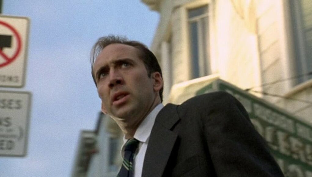 nicholas-cage-the-rock Elokuvakohtaus, jossa näyttelijä Nicolas Cage näkyy alaviistosta otetussa puolivartalokuvassa. Hänellä on yllään tumma puku, valkoinen paita ja raidallinen solmio, ja hänen ilmeensä on hämmentynyt tai järkyttynyt, suu hieman auki. Taustalla näkyy kaupunkimiljöötä, kuten vaalea rakennus, vihreä liiketilan kyltti ja vasemmassa reunassa epätarkkoja liikennemerkkejä, joissa lukee "3 tons" ja "buses". Valaistus on luonnollinen päivänvalo.