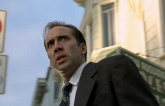 Christopher Nolan ja muut isot ohjaajat eivät suostu työskentelemään Nicholas Cagen kanssa – paljastaa syyn Elokuvakohtaus, jossa näyttelijä Nicolas Cage näkyy alaviistosta otetussa puolivartalokuvassa. Hänellä on yllään tumma puku, valkoinen paita ja raidallinen solmio, ja hänen ilmeensä on hämmentynyt tai järkyttynyt, suu hieman auki. Taustalla näkyy kaupunkimiljöötä, kuten vaalea rakennus, vihreä liiketilan kyltti ja vasemmassa reunassa epätarkkoja liikennemerkkejä, joissa lukee "3 tons" ja "buses". Valaistus on luonnollinen päivänvalo.