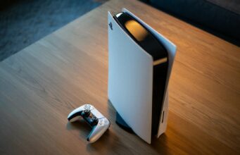 Sony ei aio myydä konsoleitaan tappiolla, mutta seuraa markkinoita PS6:n osalta Korkealaatuinen tuotekuva pystyasennossa olevasta PlayStation 5 -konsolista ja sen vieressä olevasta DualSense-ohjaimesta. Laitteet on sijoitettu vaalealle puupöydälle, ja kuva on otettu yläviistosta. Konsolin valkoinen ulkokuori ja musta keskiosa heijastavat pehmeää, lämmintä valoa, joka luo kuvaan tyylikkään ja modernin tunnelman. Tausta on osittain epätarkka, korostaen pelikonsolia kuvan keskipisteenä.
