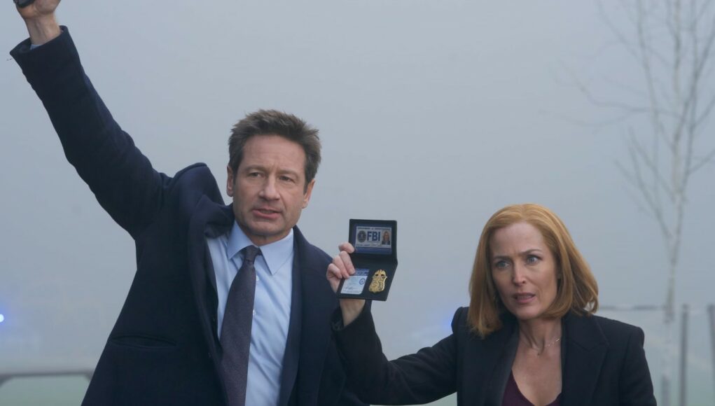 Näyttelijät David Duchovny ja Gillian Anderson rooleissaan FBI-agentteina Fox Mulderina ja Dana Scullynä. Mulder nostaa oikean kätensä ylös ja näyttää vasemmalla kädellään FBI-merkkiään. Scully seisoo hänen vieressään huolestuneen tai keskittyneen näköisenä. Molemmilla on yllään tummat puvut, ja tausta on erittäin sumuinen ja harmaa, mikä luo sarjalle tyypillisen mystisen tunnelman. Taustalla näkyy heikosti hälytysajoneuvon sinistä valoa.
