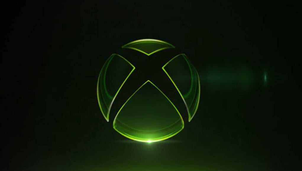 Tyylitelty Xbox-logo mustalla taustalla. Logo on läpinäkyvä ja lasimainen, ja sen ääriviivat hehkuvat kirkkaan neonvihreänä. Logon alapuolelta loistaa voimakas vihreä valonlähde, joka heijastuu peilimäiselle alustalle, ja oikealla puolella näkyy hienovarainen linssiheijastus (lens flare). Kokonaisuus on futuristinen ja minimalistinen.