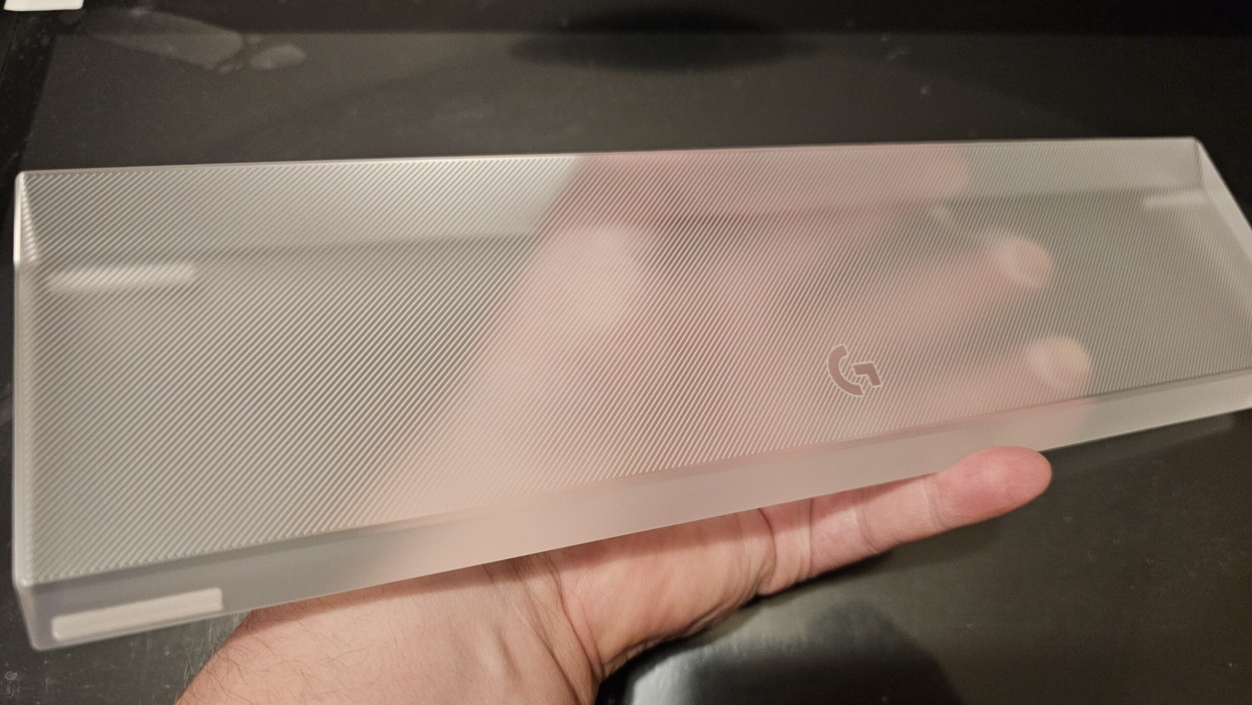 Lähikuva laserleikatusta, läpinäkyvästä akryylirannetuesta, joka lepää kämmenellä. Rannetuen pinnassa on hienon hienot, vinot urat ja sen oikeassa reunassa on kaiverrettu Logitech G -logo. Tuote näyttää kevyeltä ja sen reunat ovat kirkkaat. Taustalla näkyy epäterävästi tumma työpöytä.