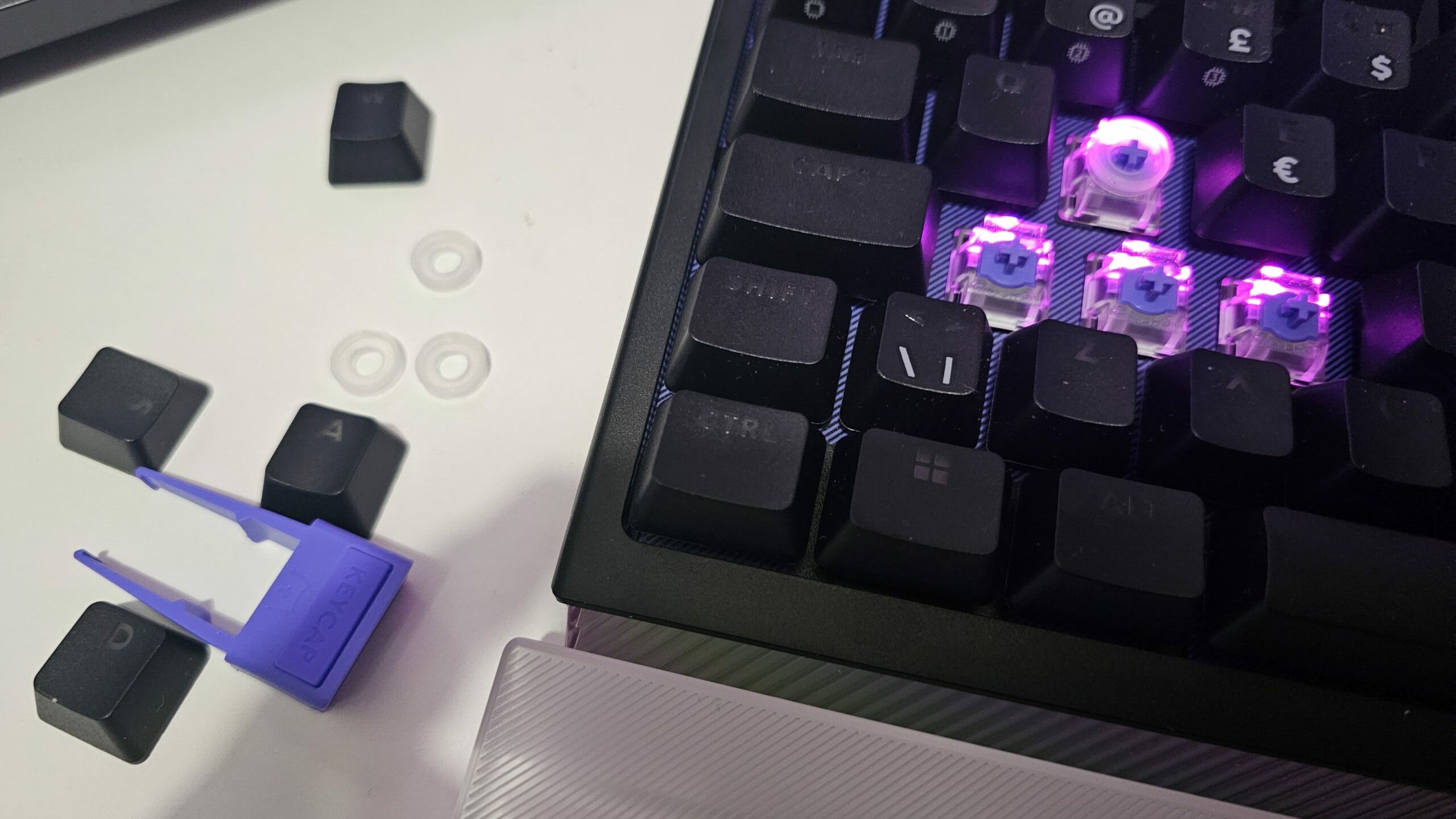 Lähikuva näppäimistöstä, jonka WASD-näppäinhatut on irrotettu. Irrotetut mustat näppäinhatut ja violetti työkalu niiden poistamiseen (keycap puller) lepäävät näppäimistön vieressä valkoisella alustalla. Niiden luona on myös kolme pientä, läpikuultavaa kumista SAPP-rengasta, jotka on tarkoitettu analogisten kytkinten kaksitoimintoa varten. Näppäimistön rungossa näkyvät paljaat kytkimet, joista yhden ympärille on asennettu tällainen rengas, ja kytkimet loistavat violettia taustavaloa.