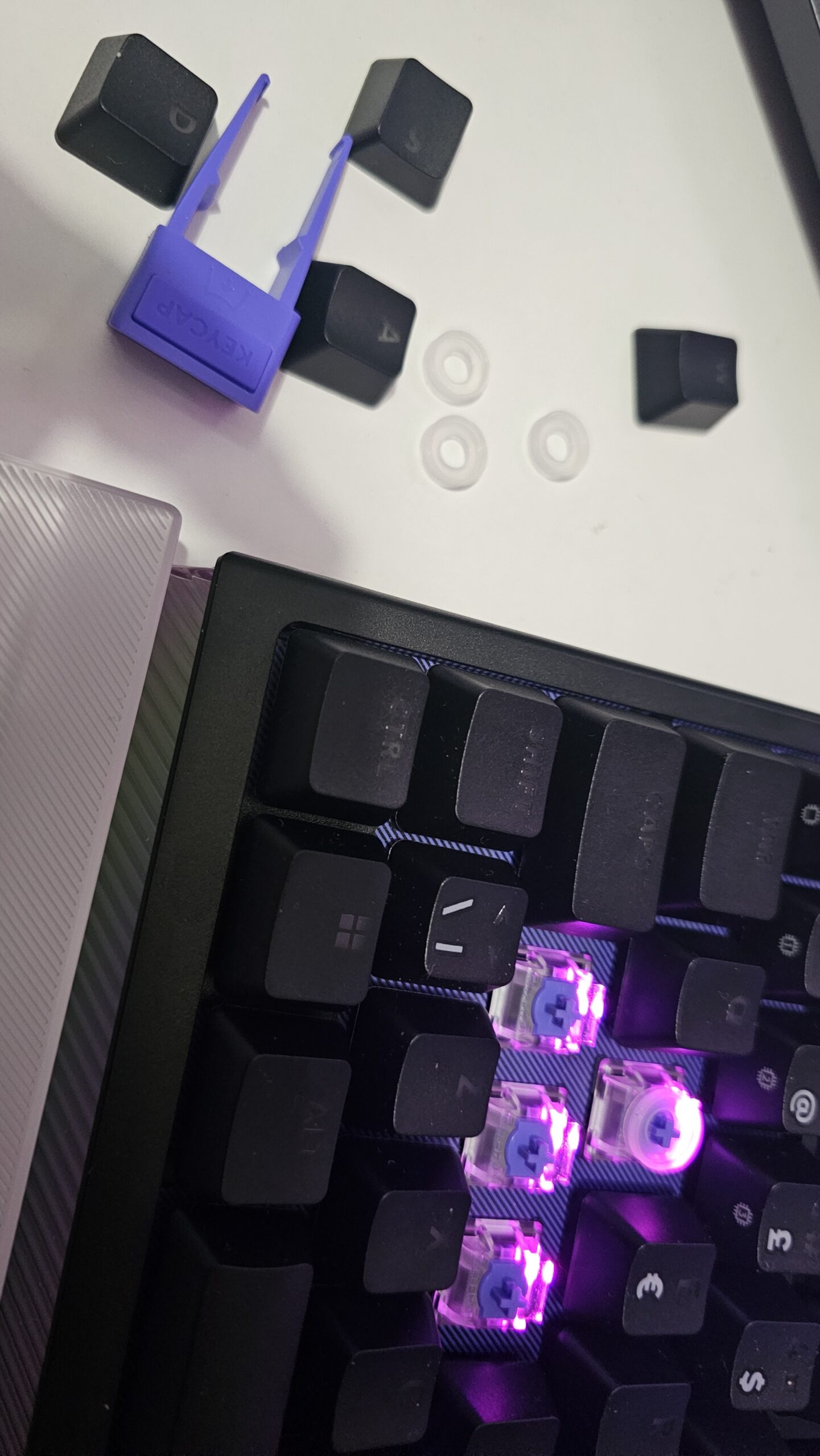 Lähikuva näppäimistöstä, jonka WASD-näppäinhatut on irrotettu. Irrotetut mustat näppäinhatut ja violetti työkalu niiden poistamiseen (keycap puller) lepäävät näppäimistön vieressä valkoisella alustalla. Niiden luona on myös kolme pientä, läpikuultavaa kumista SAPP-rengasta, jotka on tarkoitettu analogisten kytkinten kaksitoimintoa varten. Näppäimistön rungossa näkyvät paljaat kytkimet, joista yhden ympärille on asennettu tällainen rengas, ja kytkimet loistavat violettia taustavaloa.