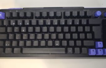 Logitech G512 X 75 -pelinäppäimistön arvostelu – tee-se-itse-Frankenspelinäppis Ylhäältä päin kuvattu kompakti, musta mekaaninen pelinäppäimistö valkoisella alustalla. Näppäimistössä on 75 prosentin asettelu ja mustat näppäinhatut, joita koristavat violetit tehosteet: vasemman yläkulman Esc-näppäin Logitech G -logolla, oikean alakulman nuolinäppäimet sekä oikeassa yläkulmassa olevat kaksi pyöreää rullasäädintä. Laitteen pinnassa näkyy käytön jälkiä ja hienojakoista pölyä.