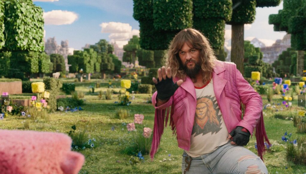 jason-momoa-minecraft-2025 Elokuvakohtaus Minecraft-teemaisesta maailmasta, jossa näyttelijä Jason Momoa esiintyy pitkähiuksisena ja parrakkaana hahmona. Hänellä on yllään erottuva, hapsullinen vaaleanpunainen nahkatakki, graafinen t-paita leijonakuvioinnilla sekä revityt vaaleat farkut. Hän on kyykistynyt tai astunut askeleen eteenpäin vehreällä, kuutiomaisella ruohokentällä ja nostanut sormikkaalla suojatun oikean kätensä eteenpäin kämmen edellä, aivan kuin varoittaakseen jotakuta tai käyttäen taikaa. Vasemmassa etukulmassa näkyy sumea, vaaleanpunainen olento (mahdollisesti sika). Taustalla avautuu aurinkoinen, palikoista koostuva Minecraft-maisema, jossa on neliömäisiä puita, kukkia ja kuutiomaisia pilviä sinisellä taivaalla.