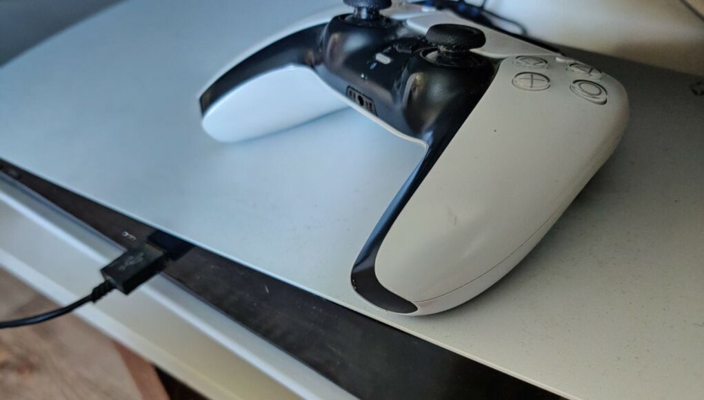 Lähikuva PlayStation 5 -konsolin päällä lepäävästä valkoisesta DualSense-ohjaimesta, kun konsoliin on kytkettynä musta USB-kaapeli ja laitteen alaosasta heijastuu lämmin valo tasolle.