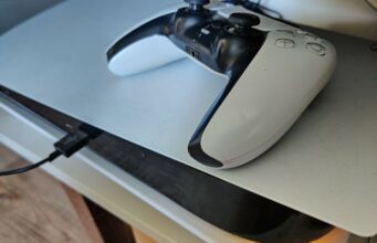 Nyt se on päätetty: Sony ei enää julkaise omia pelejään PC:lle tai muille alustoille Lähikuva PlayStation 5 -konsolin päällä lepäävästä valkoisesta DualSense-ohjaimesta, kun konsoliin on kytkettynä musta USB-kaapeli ja laitteen alaosasta heijastuu lämmin valo tasolle.