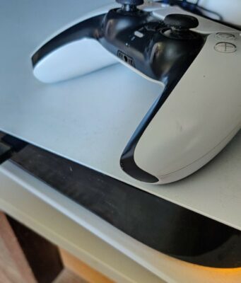 Sony laittoi oudot varoitustarrat uusiin PS5-konsolipakkauksiinsa Lähikuva PlayStation 5 -konsolin päällä lepäävästä valkoisesta DualSense-ohjaimesta, kun konsoliin on kytkettynä musta USB-kaapeli ja laitteen alaosasta heijastuu lämmin valo tasolle.