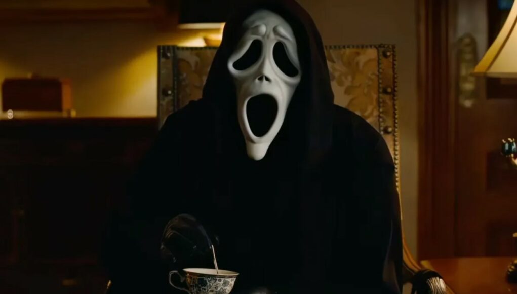 scary-movie-6 Scary Movie -elokuvista tuttu parodia-Ghostface-hahmo istuu mustassa kaavussaan ja naamiossaan nojatuolissa sekoittaen lusikalla teetä kuvioidussa teekupissa hämärästi valaistussa huoneessa.
