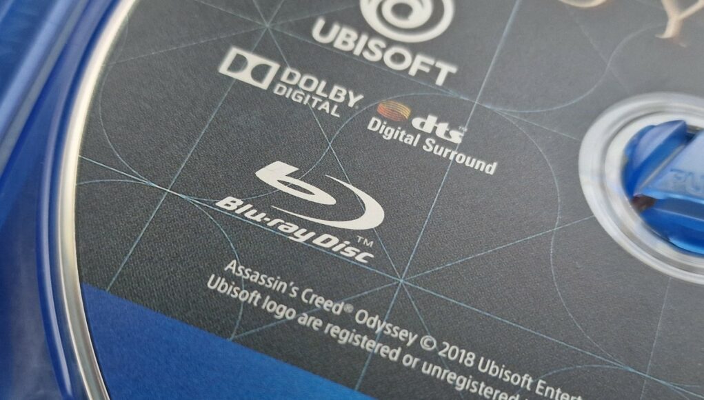 fyysinen-assassins-creed-odyssey-pelilevy-lahikuva Lähikuva Assassin's Creed Odyssey -pelilevyn painatuksesta, jossa näkyvät Blu-ray Disc -logo sekä ääni- ja julkaisijatiedot.