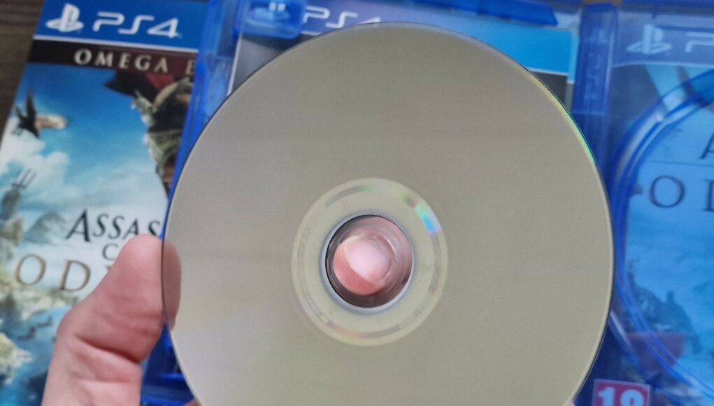 fyysinen-pelilevy Käsi pitelee PlayStation 4 -pelin levyä, jonka taustalla näkyy pelikotelo.