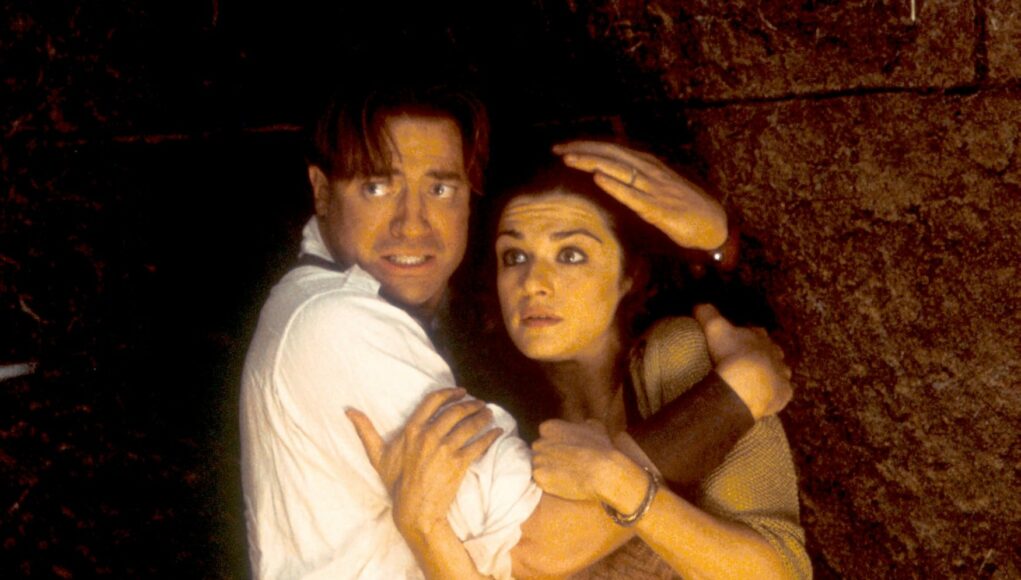 Brendan Fraser ja Rachel Weisz pelästyneinä ja toisiinsa kietoutuneina hämärässä ja karussa ympäristössä.